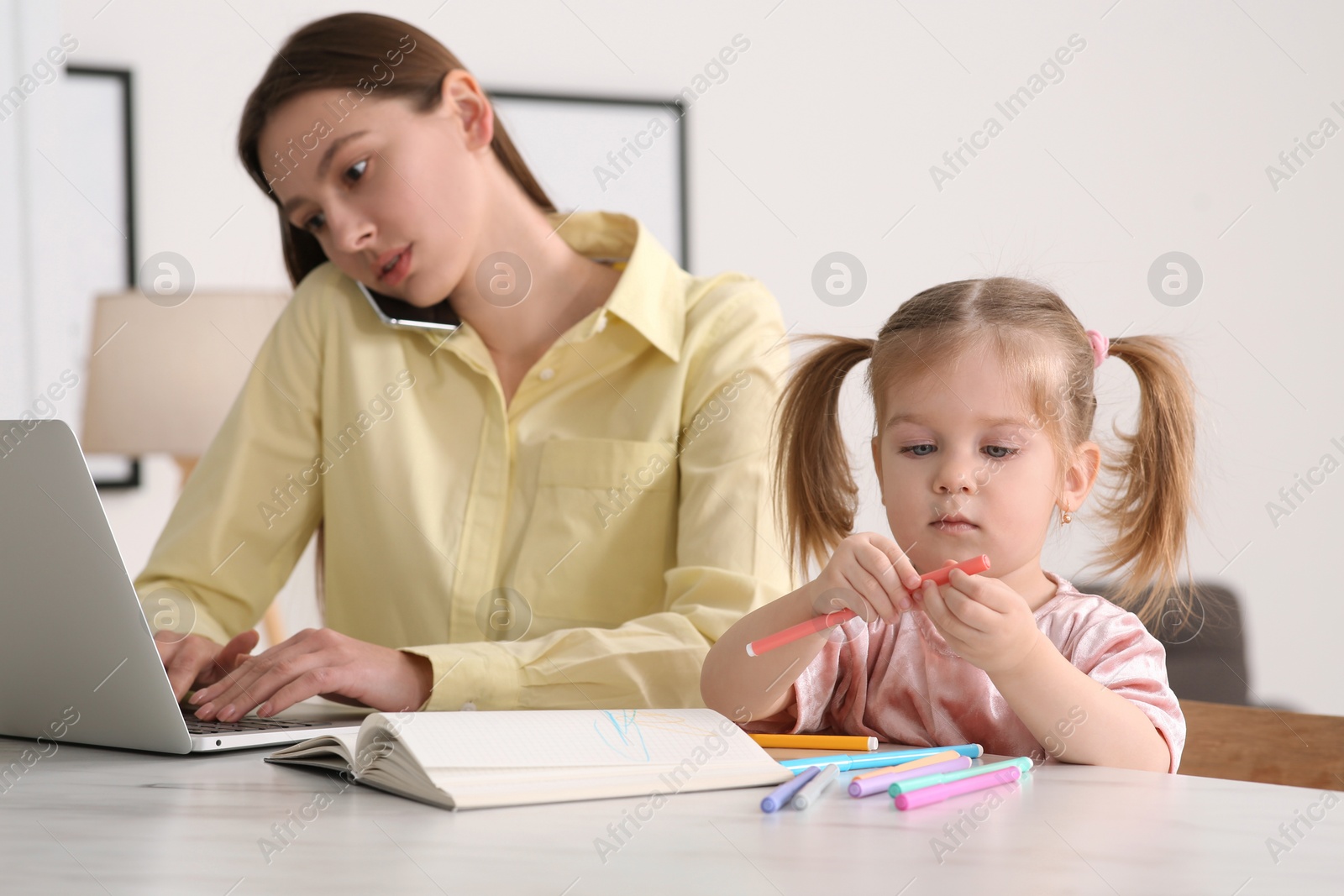 Photo of Woman with laptop working remotely at home. Busy mother talking on phone while her daughter drawing at desk. Selective focus