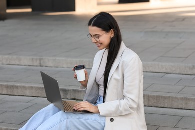 Happy young woman with cup of coffee using modern laptop on stairs outdoors