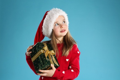 Photo of Cute child in Santa hat with Christmas gift on light blue background