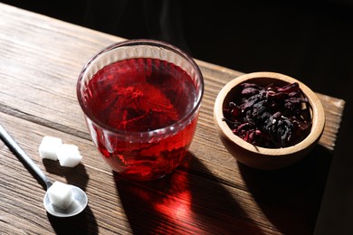 Delicious hibiscus tea in glass, sugar cubes and dry roselle petals on wooden table, closeup
