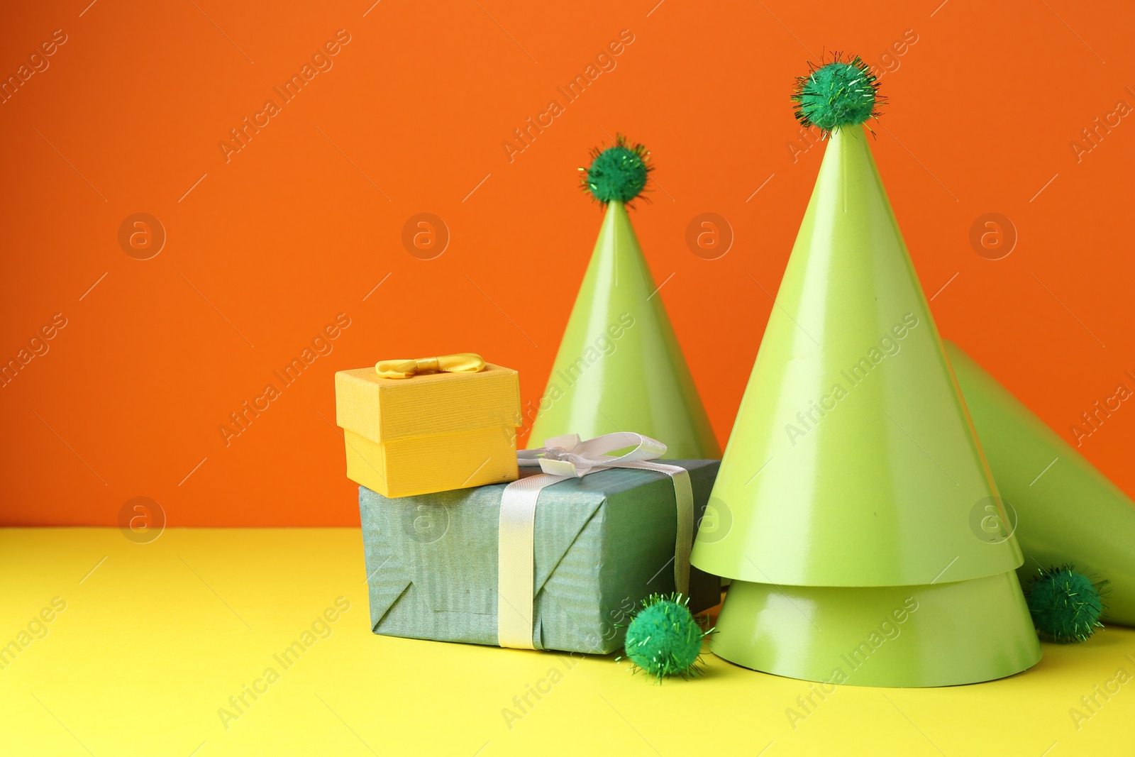 Photo of Party hats and gift boxes on yellow table against orange background, space for text