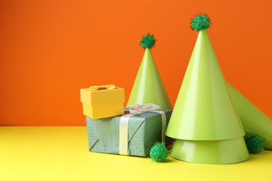 Party hats and gift boxes on yellow table against orange background, space for text