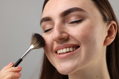 Smiling woman with freckles applying makeup with brush on grey background, closeup