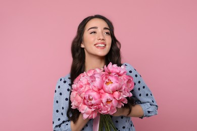 Beautiful young woman with bouquet of peonies on pink background