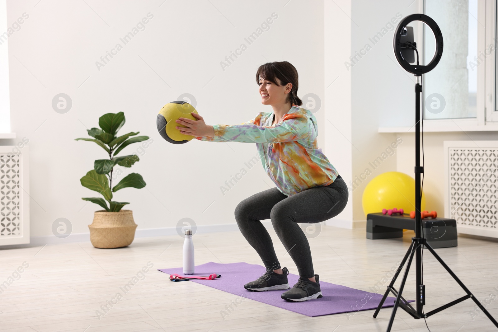 Photo of Happy sports blogger training with medicine ball while streaming online fitness lesson with smartphone at home