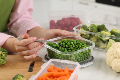 Woman taking green peas with spoon from glass container at white marble table in kitchen, closeup. Food storage