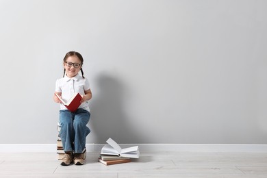 Cute little girl in glasses sitting on stack of books near light grey wall. Space for text