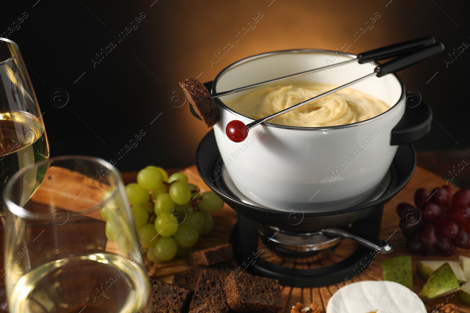 Photo of Forks with pieces of grape, bread, fondue pot with melted cheese, wine and snacks on table, closeup