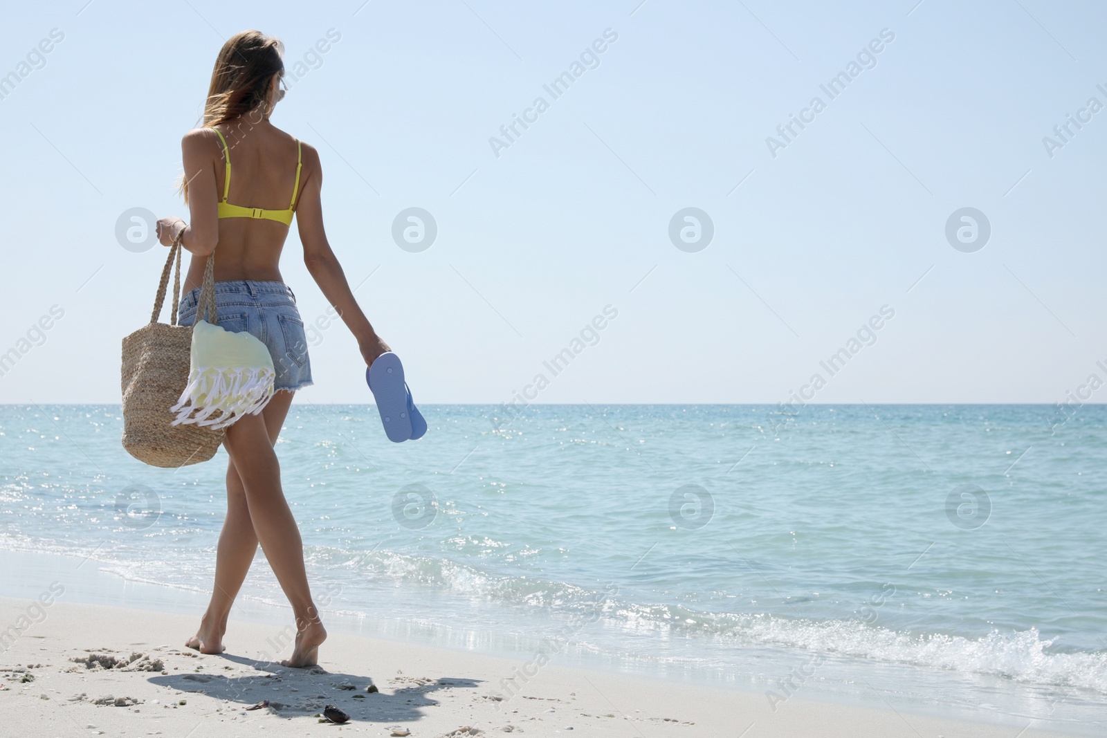 Photo of Beautiful woman with beach bag and flip flops walking near sea, back view