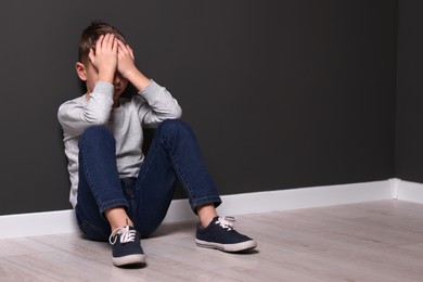 Photo of Child abuse. Upset boy sitting on floor near gray wall indoors, space for text
