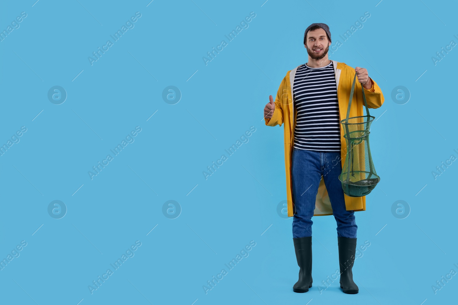 Photo of Fisherman holding fishing net with catch and showing thumb up on light blue background, space for text