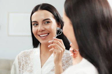 Photo of Beautiful young woman with eyelash curler near mirror indoors