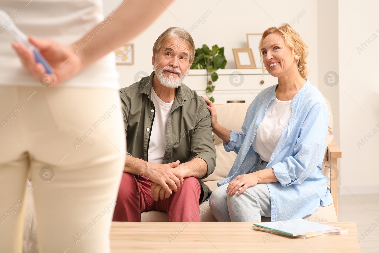 Photo of Woman holding pregnancy test behind her back near parents indoors. Grandparents' reaction to future grandson