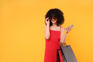 Happy young woman with shopping bags and modern smartphone on yellow background. Space for text