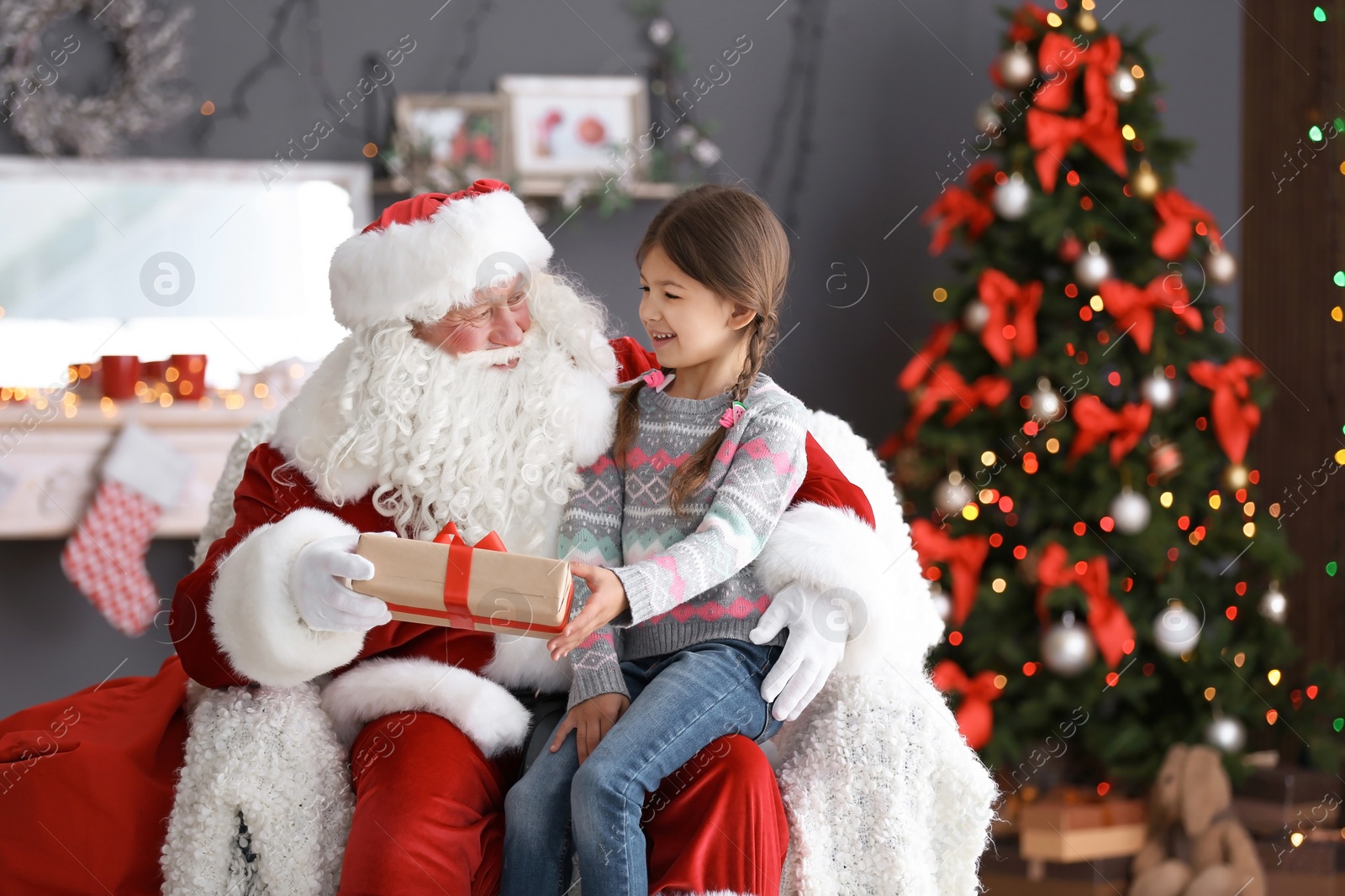 Photo of Little girl with gift box sitting on authentic Santa Claus' lap indoors