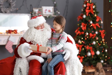Photo of Little girl with gift box sitting on authentic Santa Claus' lap indoors