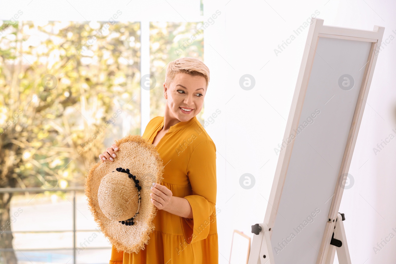 Photo of Beautiful mature woman with hat looking at herself in large mirror indoors