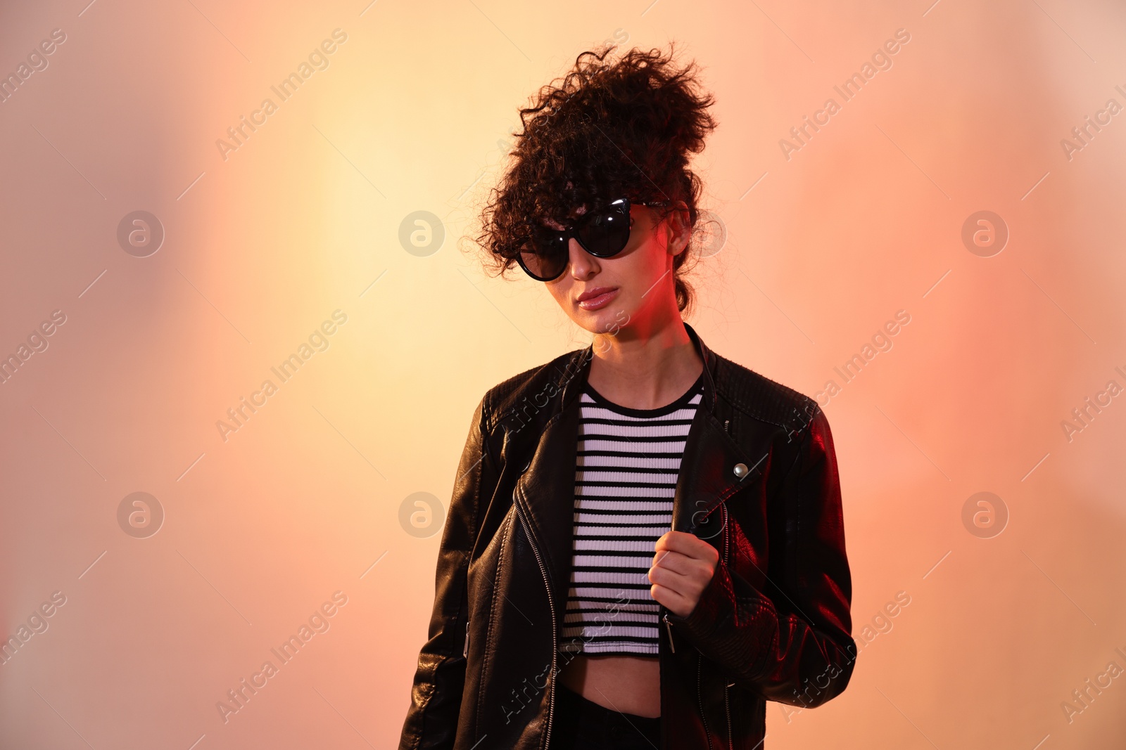 Photo of Beautiful young woman in black leather jacket and sunglasses posing on color background in neon lights