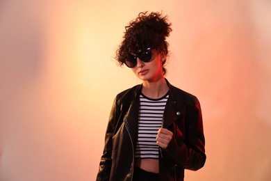 Photo of Beautiful young woman in black leather jacket and sunglasses posing on color background in neon lights