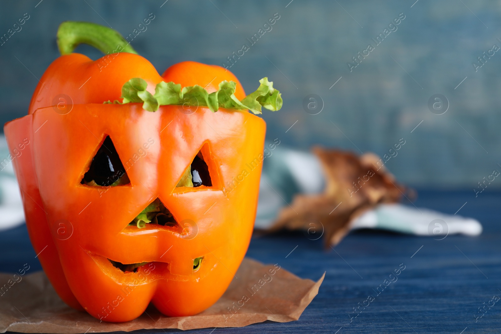 Photo of Bell pepper with black olives and lettuce as Halloween monster on blue wooden table. Space for text