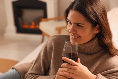 Happy young woman with glass of wine relaxing near fireplace at home. Space for text