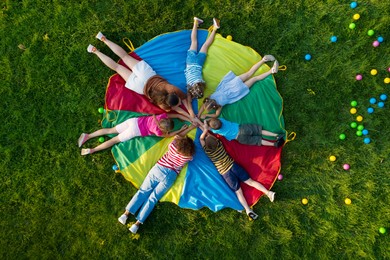 Image of Group of children with teachers holding hands together on rainbow playground parachute in park, top view. Summer camp activity