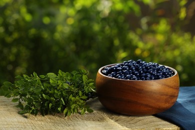 Delicious bilberries in bowl and branch with fresh berries on wooden table outdoors