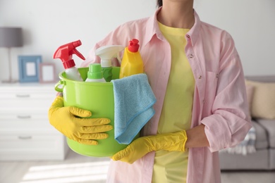 Photo of Woman holding bucket with different cleaning supplies at home, closeup