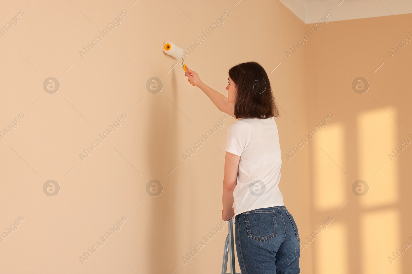 Photo of Young woman painting wall with roller on stepladder indoors, space for text. Room renovation