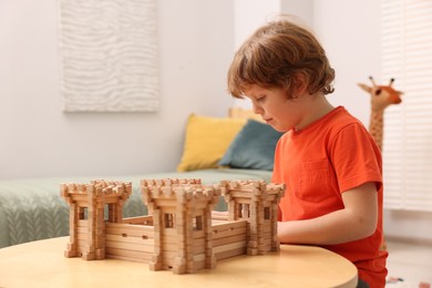 Cute little boy playing with wooden fortress at table in room. Child's toy