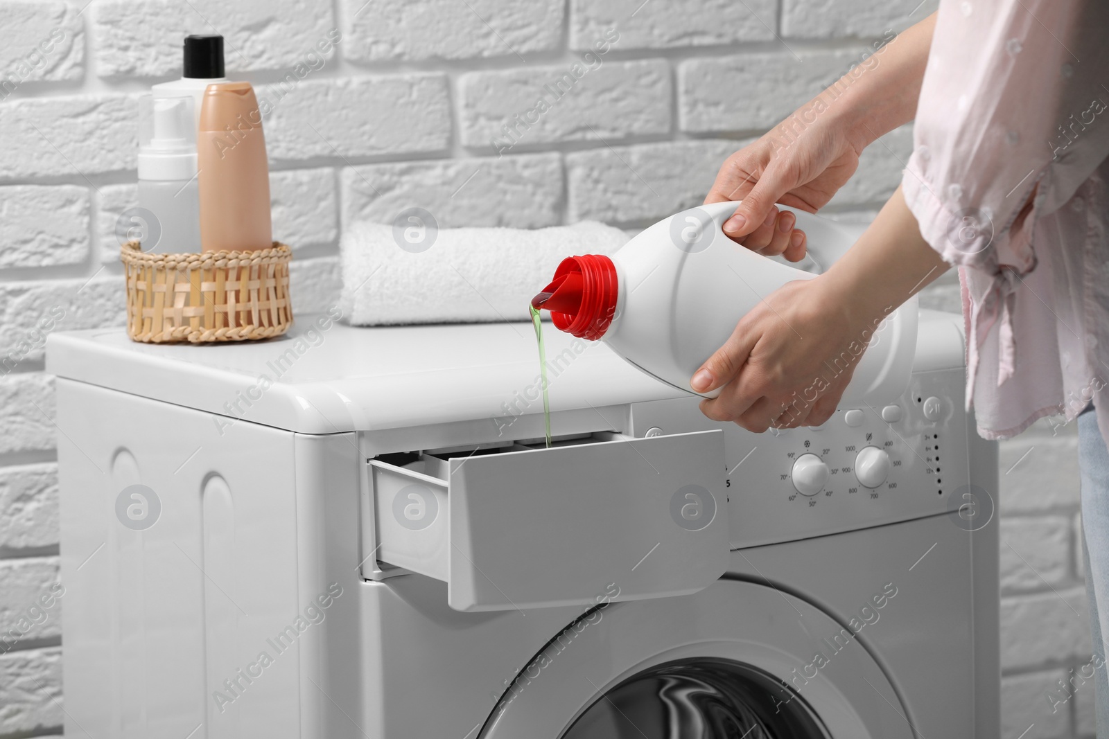 Photo of Woman pouring fabric softener from bottle into washing machine near white brick wall, closeup