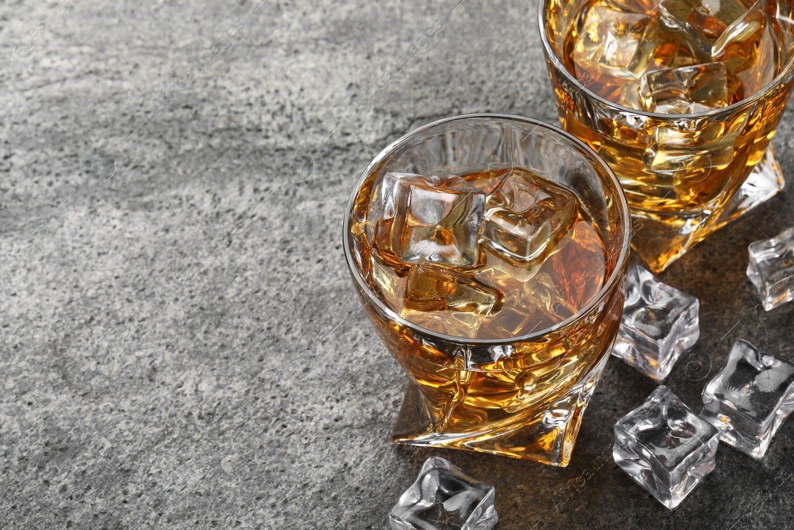 Photo of Whiskey and ice cubes in glasses on grey textured table, space for text