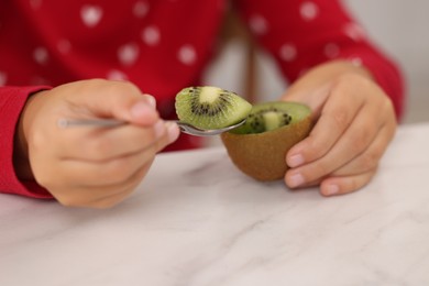 Photo of Girl eating tasty fresh kiwi with spoon at white marble table indoors, closeup