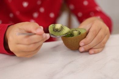 Girl eating tasty fresh kiwi with spoon at white marble table indoors, closeup