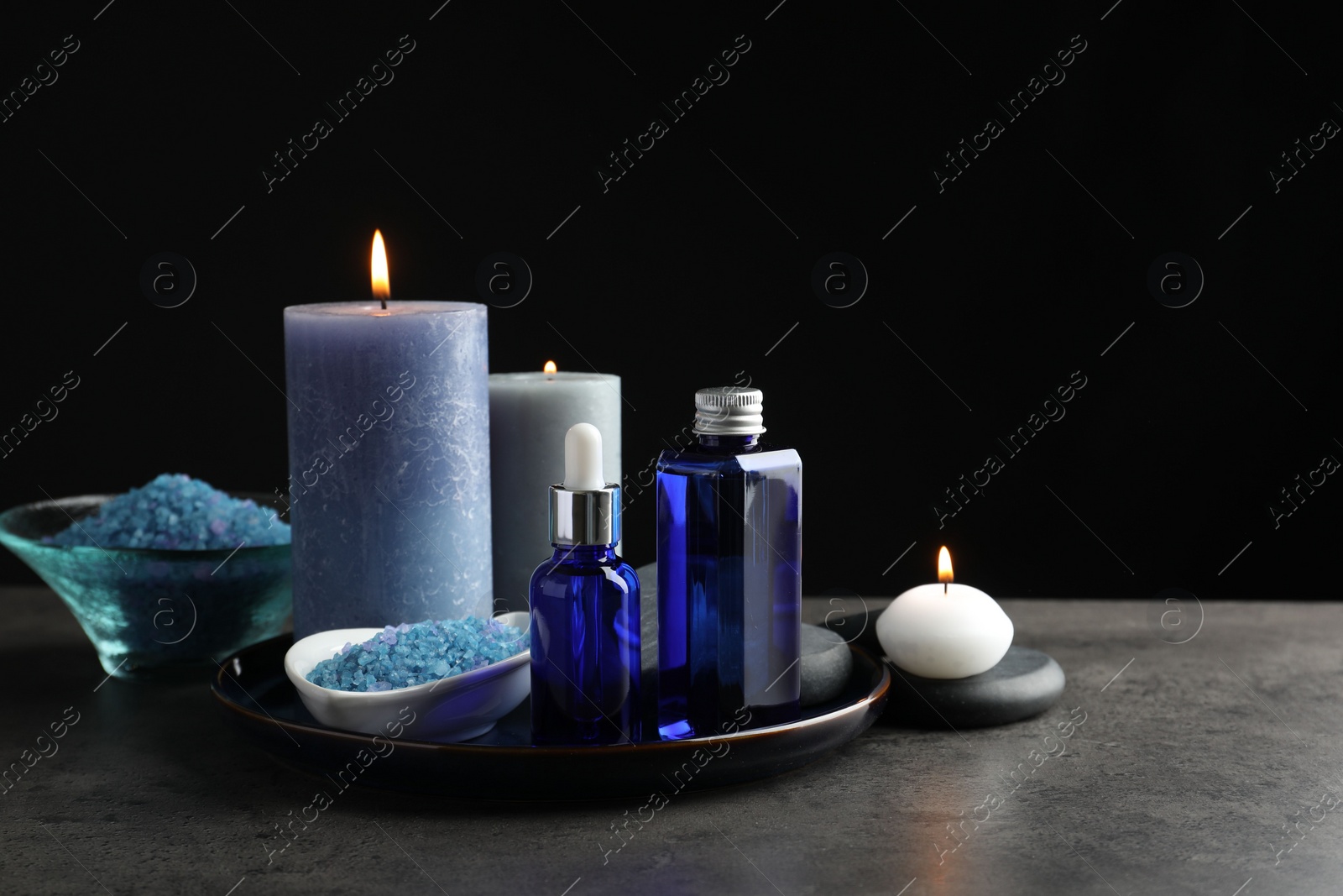 Photo of Spa composition. Cosmetic products, burning candles and sea salt on gray table against black background, space for text