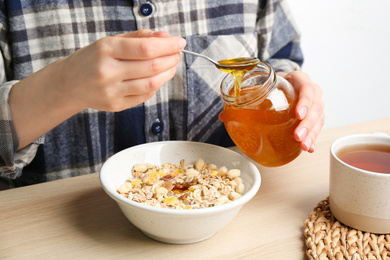 Photo of Woman putting honey into bowl with muesli at wooden table, closeup