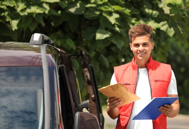 Photo of Young courier with clipboard and envelope near delivery car outdoors