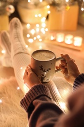 Photo of Woman with cup of drink on floor against blurred Christmas lights, closeup