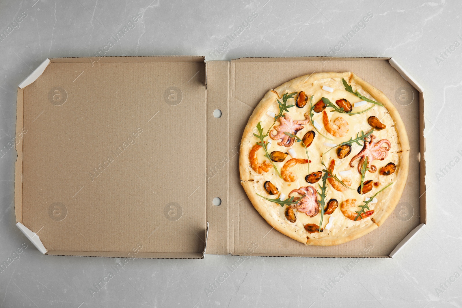 Photo of Delicious seafood pizza in cardboard box on grey marble table, top view