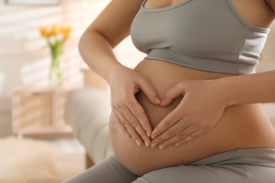Photo of Pregnant young woman making heart with her hands on belly at home, closeup. Space for text