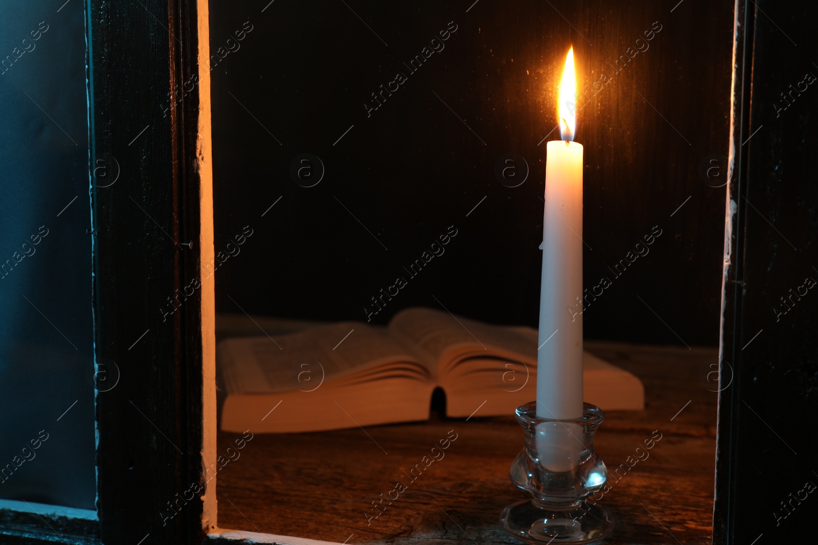 Photo of Burning candle and Bible on wooden table at night, view through window