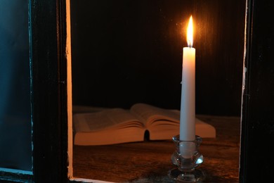 Photo of Burning candle and Bible on wooden table at night, view through window