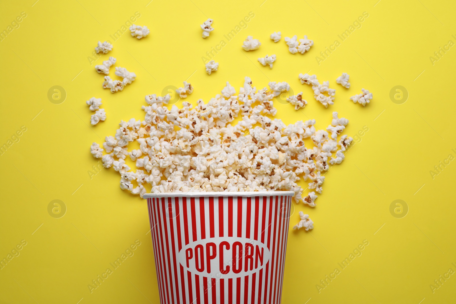 Photo of Overturned paper bucket with delicious popcorn on yellow background, flat lay