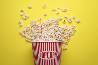 Overturned paper bucket with delicious popcorn on yellow background, flat lay