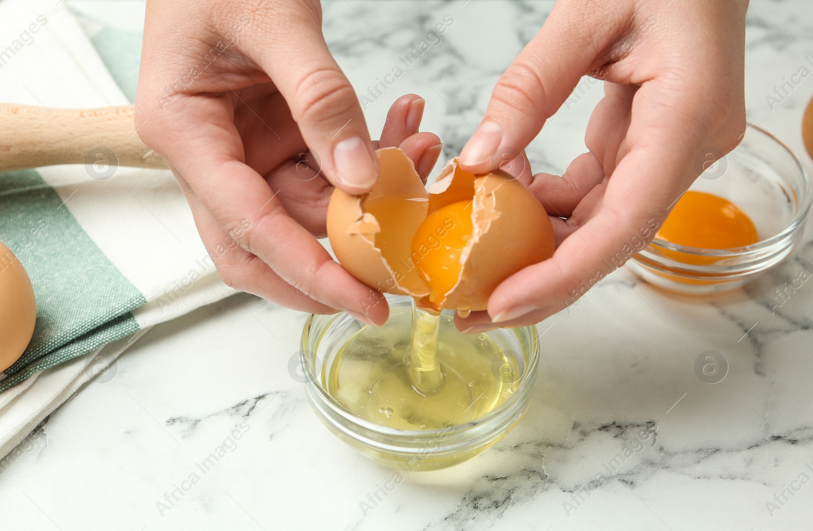 Photo of Woman separating egg yolk from white over glass bowl at marble table, closeup