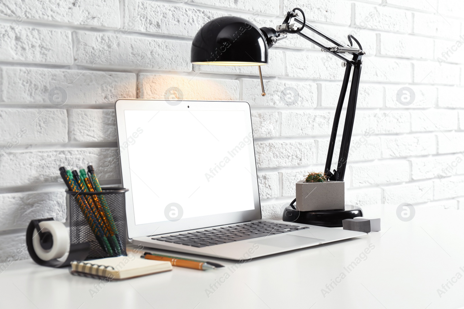 Photo of Modern workplace with laptop and lamp on table near brick wall. Space for design