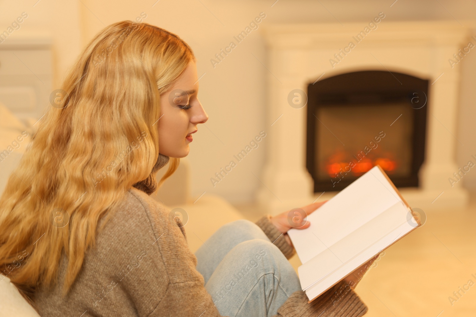 Photo of Beautiful young woman reading book near fireplace in room