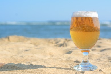 Glass of cold beer on sandy beach near sea. Space for text