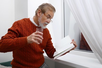 Photo of Upset senior man with glass of water reading book near window at home. Loneliness concept