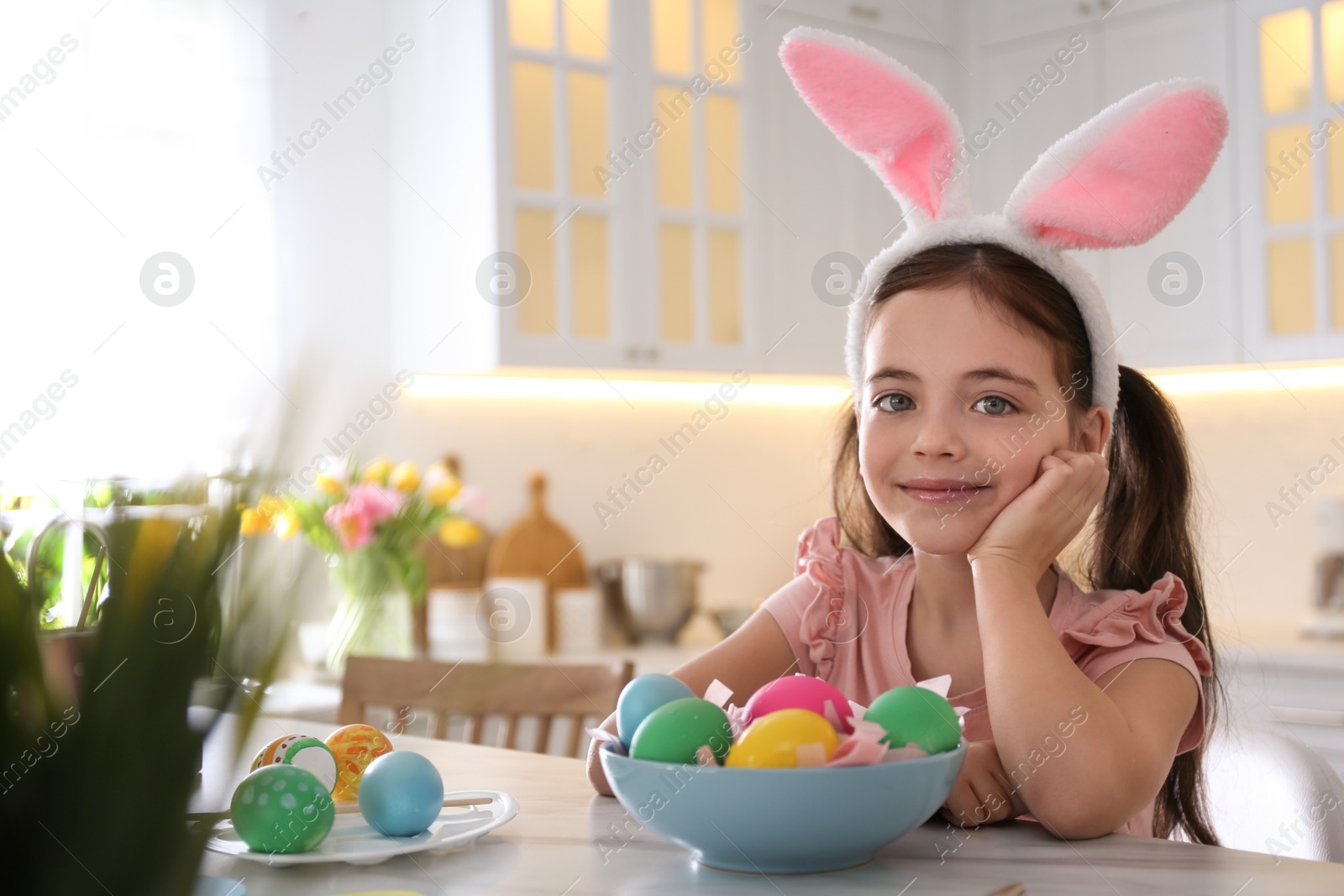 Photo of Cute little girl in bunny ears headband painting Easter eggs at table indoors